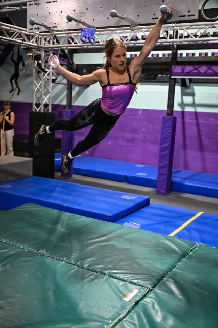 An athlete swinging from an obstacle above purple mats