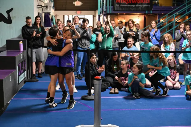 Two athletes hugging on the course as a crowd of girls and parents cheers behind them