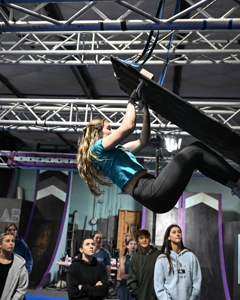 An athlete in a teal shirt hanging from the handle of a tilted board obstacle as a crowd of women and girls watches at the WNA x MLAB event