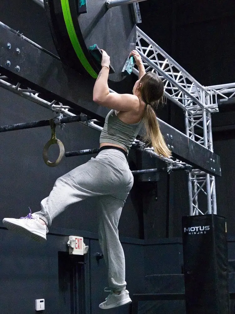 An athlete in grey sweatpants pulling up on the handles of a big wheel obstacle on the Motus Ninjas course