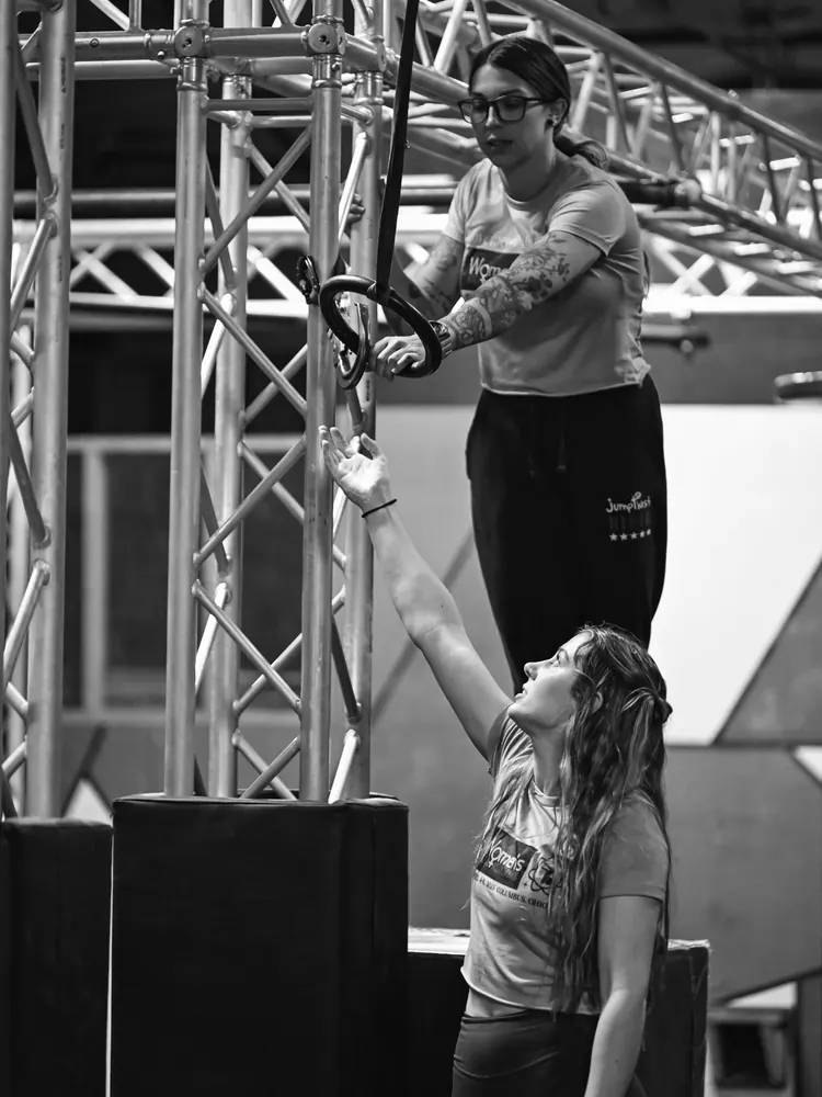 In black and white, a woman in a WNA shirt hands a gymnastic ring down from the course to an athlete reaching up for it