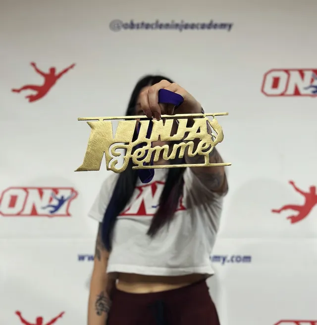 A gold Ninja Femme medal held up in front of the Obstacle Ninja Academy backdrop