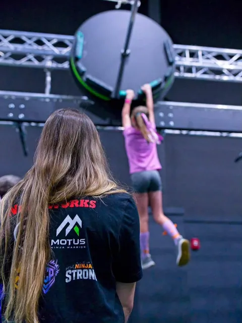 Seen from behind a coach in a Motus shirt, a young athlete in pink hangs from the handles of a big spinning wheel obstacle