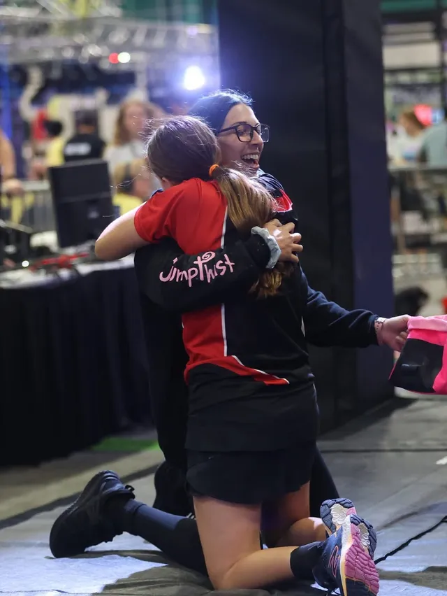 A coach kneeling on the mat to hug a young athlete in a Jumptwist jersey