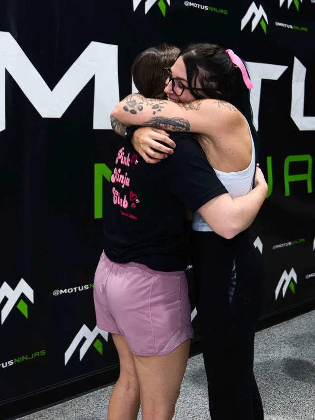 Two athletes hugging in front of the Motus Ninjas banner at the Season 1 Finals, one in a Pink Ninja Club shirt